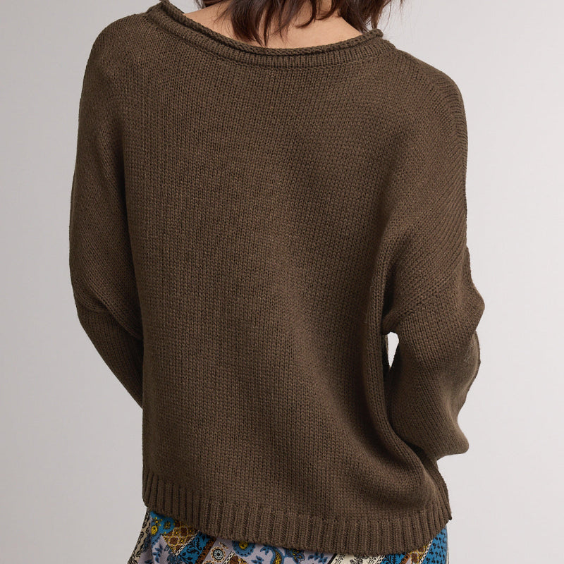 A woman viewed from the back wearing a dark brown ribbed knit sweater and a patterned skirt, against a plain light gray studio background.