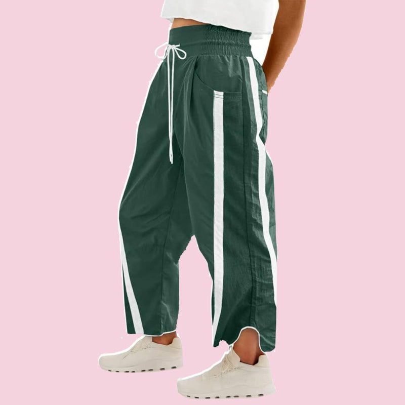 A person wearing dark green wide-leg track pants with white side stripes and a white drawstring waist, paired with white sneakers against a solid pink background.