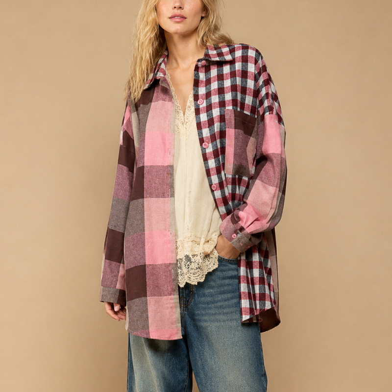 A woman stands against a plain beige studio background wearing an oversized pink and red plaid button-up shirt over a cream lace-trim blouse, wide-leg blue jeans, and brown shoes.