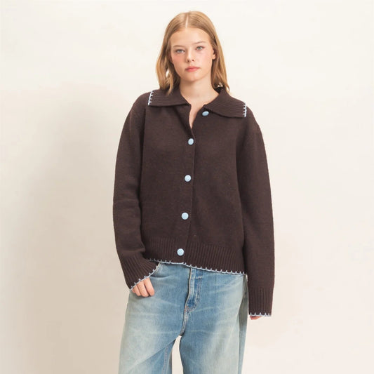 A woman modeling a dark brown knit button-up cardigan with light blue buttons and white trim, paired with wide-leg light blue jeans and beige shoes, against a plain off-white studio background.
