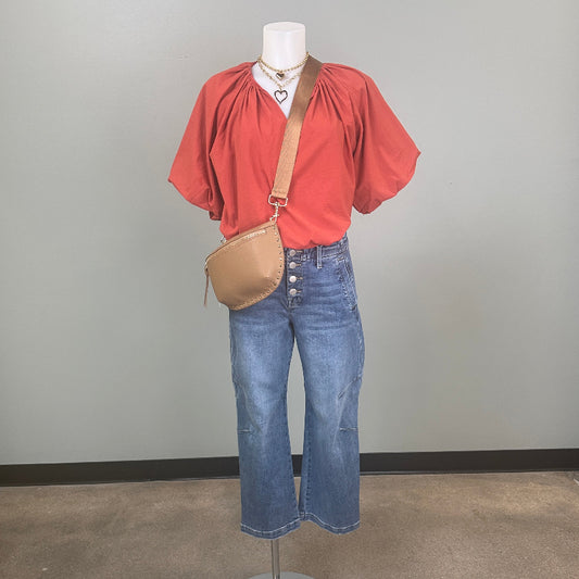 Mannequin wearing a orange top and blue jeans with a beige bag in a minimal indoor setting.