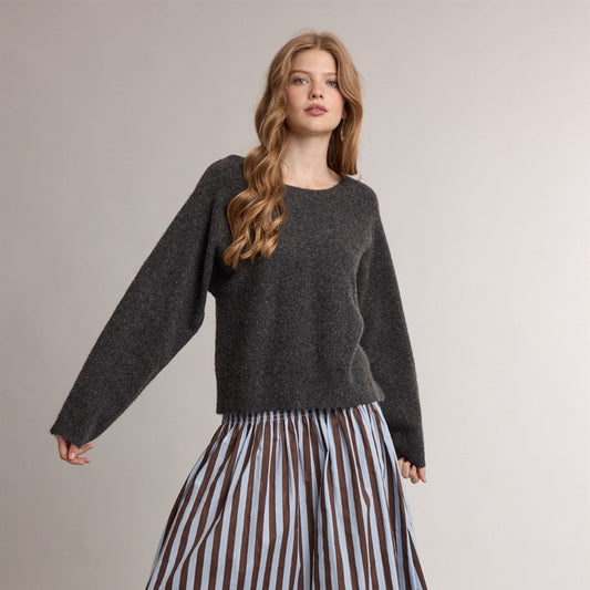 A woman modeling a dark gray knit sweater and a pleated midi skirt with vertical brown, white, and light blue stripes, paired with dark brown loafers, against a plain light gray studio background.
