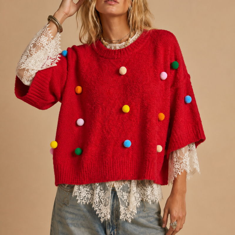A woman models a bright red knit sweater featuring colorful pom-pom accents and white lace trim on the sleeves and hem, paired with light-wash jeans against a solid beige background.