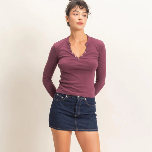 A woman modeling a long-sleeve henley top in dusty mauve-purple with a scalloped neckline, paired with a dark blue denim mini skirt and black flats, against a plain off-white studio background.