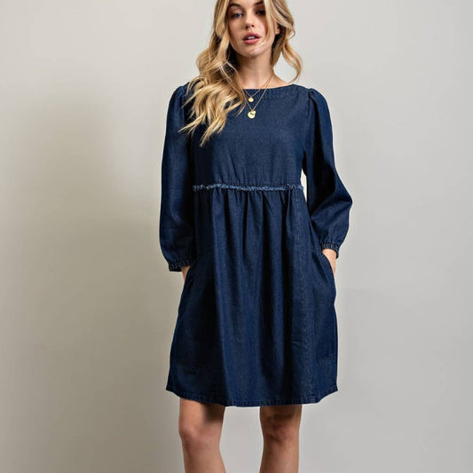 A woman modeling a dark blue long-sleeve dress with a gathered waist and side pockets, paired with beige strappy high-heeled sandals, against a plain light gray studio background.