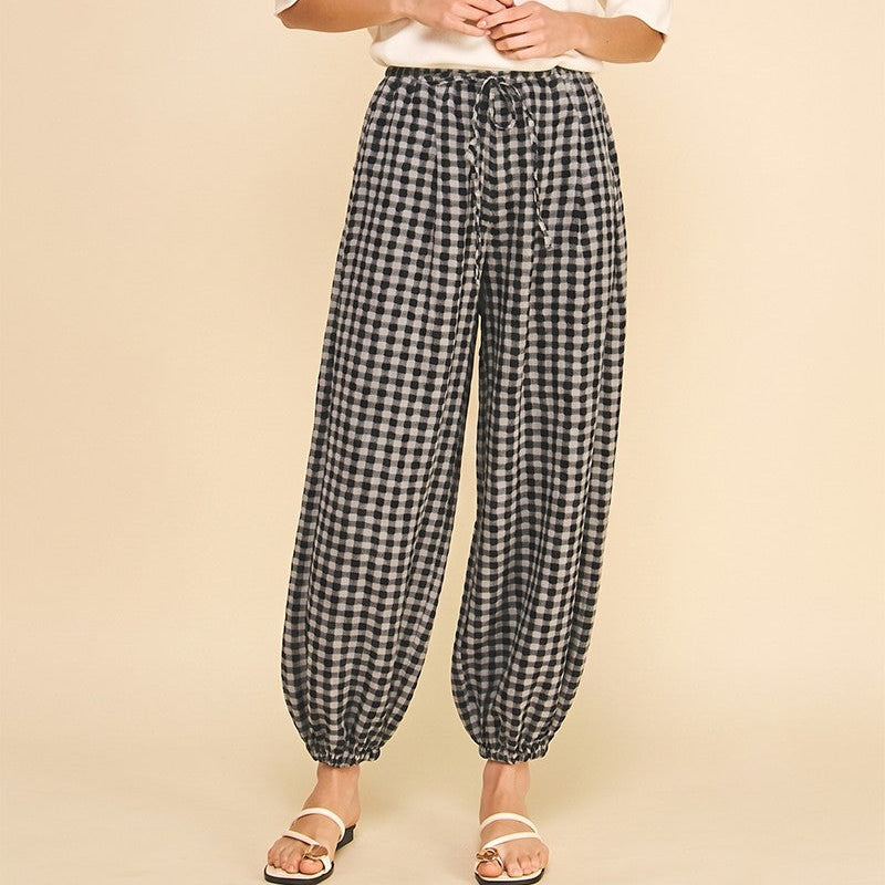 A woman modeling black and white gingham wide-leg pants with a drawstring waist, paired with a white t-shirt and flat sandals, against a solid beige studio background.