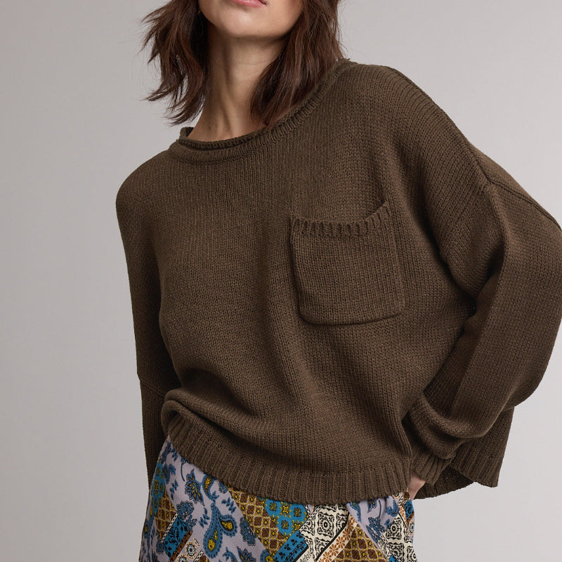 A woman wearing an oversized brown knit sweater with a chest pocket and a patterned skirt, photographed against a plain light gray studio background.