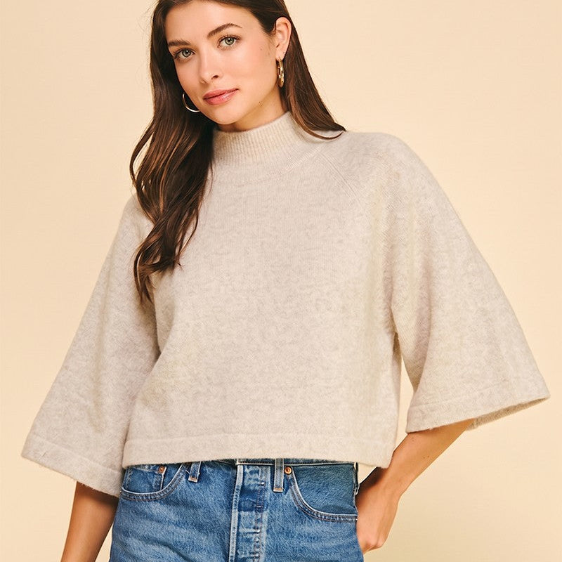 A woman modeling a light beige knit mock-neck poncho sweater with wide flared sleeves, paired with blue denim shorts, against a solid beige background.