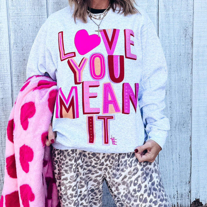 Person wearing a sweatshirt with 'LOVE YOU MEAN IT' text, holding a pink blanket with heart patterns against a wooden background.