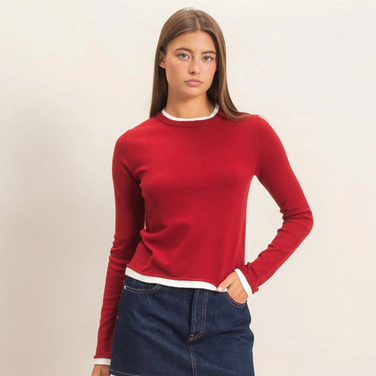 A woman modeling a red long-sleeve crewneck sweater with white ribbed trim, paired with a dark blue denim mini skirt and black flats, against a plain light beige studio background.