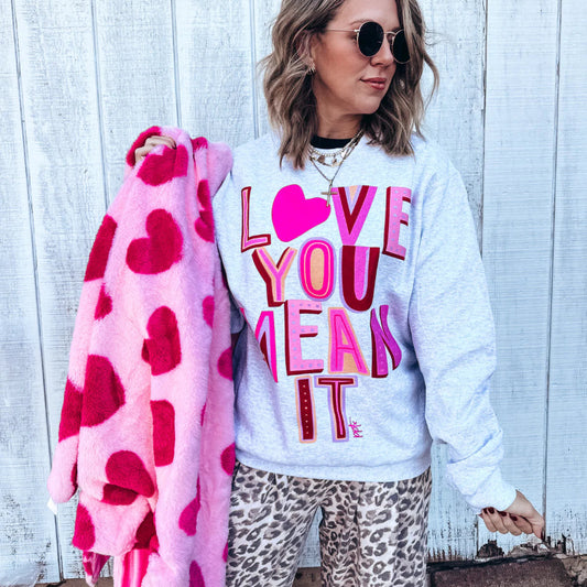 Woman wearing a white sweatshirt with colorful text, holding a pink blanket with heart patterns, standing against a light wooden wall.