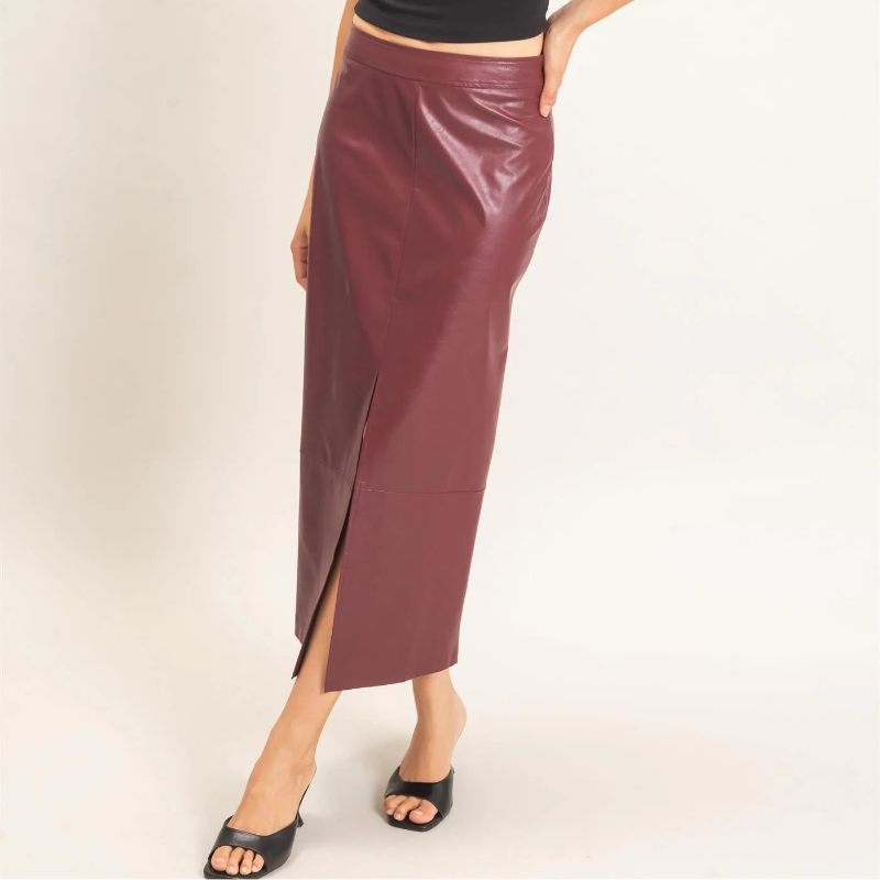 A woman modeling a black short-sleeve crop top and a shiny burgundy midi skirt with a side slit, paired with black slide sandals, against a plain off-white studio background.