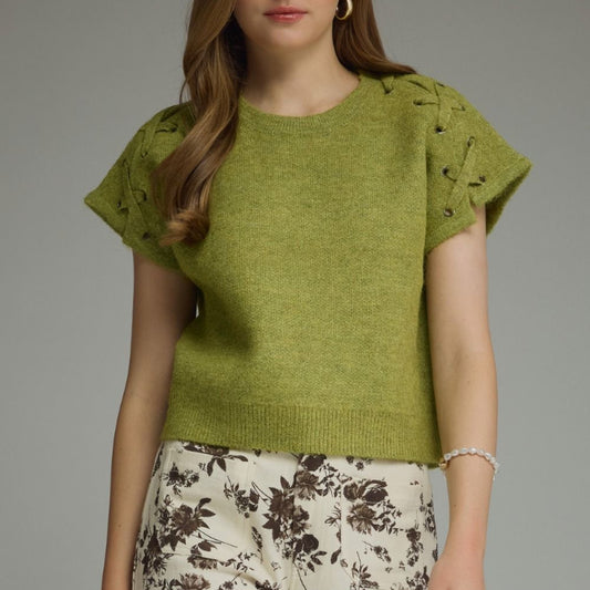 A woman modeling a short-sleeve knit top in olive green with textured, openwork shoulder details, paired with cream floral pants, against a solid gray studio background.