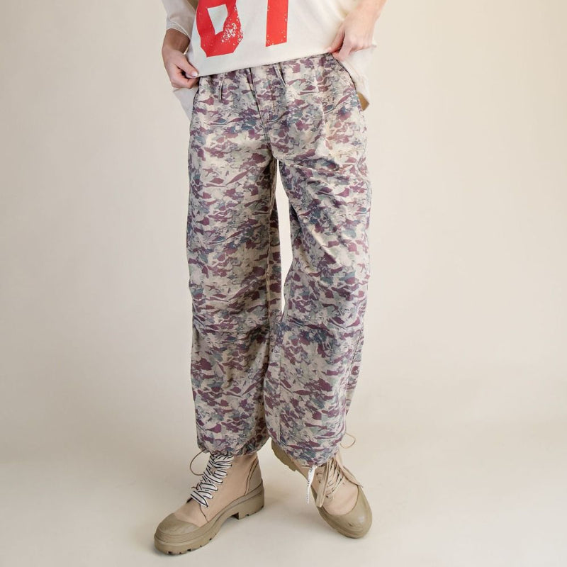 A woman modeling a cream sweatshirt with a large red '67' graphic, paired with loose-fit floral camouflage pants and beige combat boots, against a solid beige studio background.