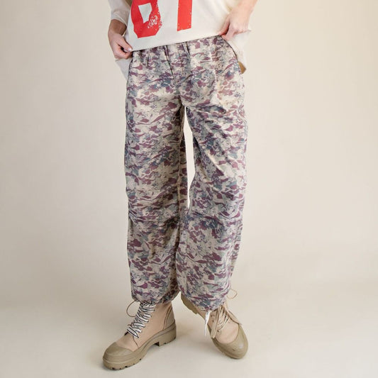 A woman modeling a cream sweatshirt with a large red '67' graphic, paired with loose-fit floral camouflage pants and beige combat boots, against a solid beige studio background.