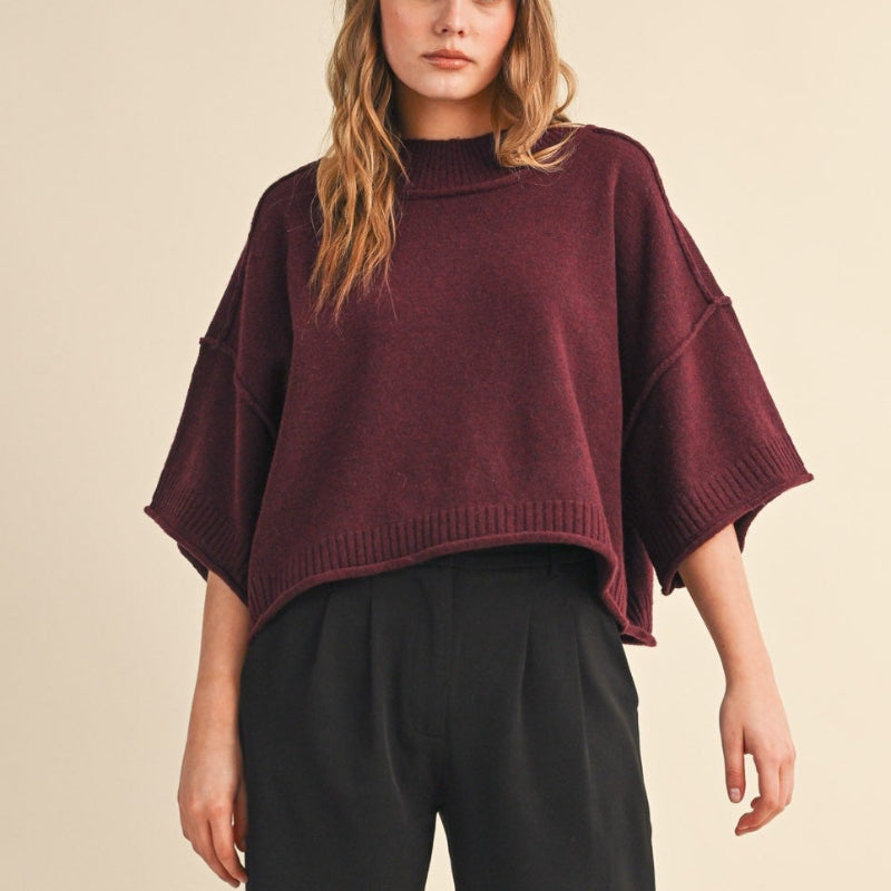 A model stands against a plain beige studio background wearing a cropped burgundy knit sweater with wide short sleeves, black wide-leg trousers, black shoes, and a cream beret.