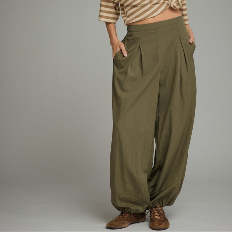A woman modeling a beige and brown horizontally striped crop top with three-quarter sleeves, paired with olive green wide-leg pants and brown shoes, against a solid gray studio background.
