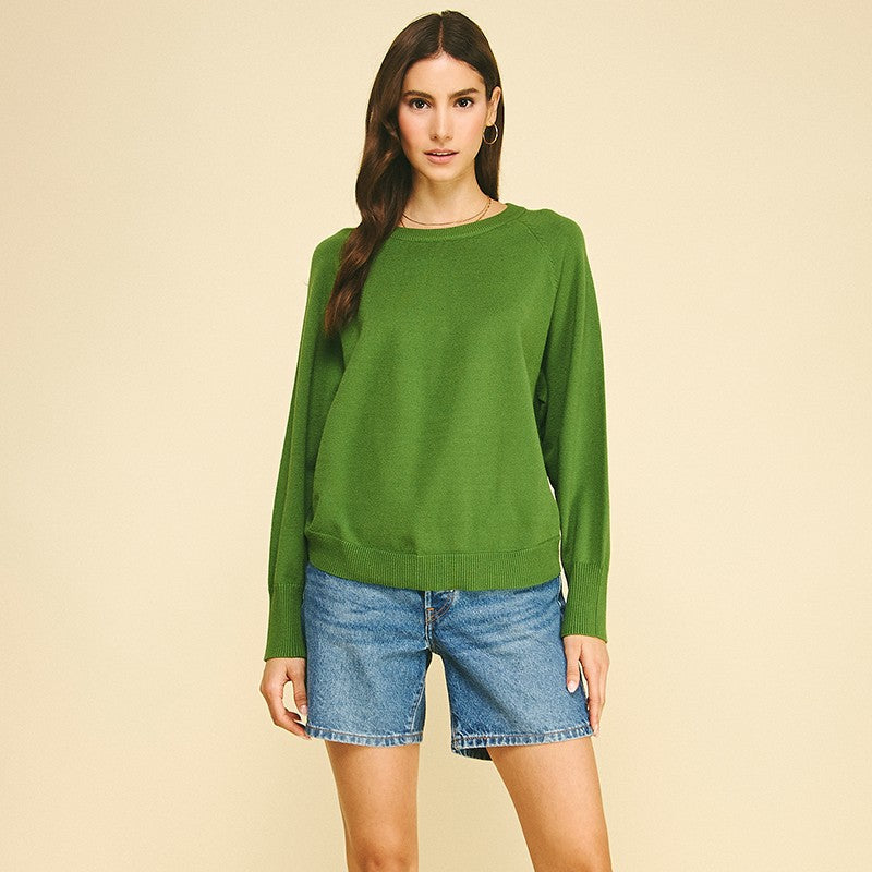 A woman modeling a green long-sleeve crewneck sweater, paired with denim shorts and tan suede boots, against a solid beige studio background.