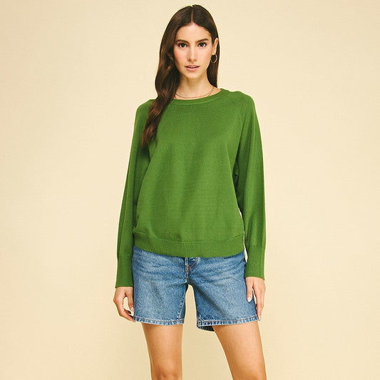 A woman modeling a green long-sleeve crewneck sweater, paired with denim shorts and tan suede boots, against a solid beige studio background.
