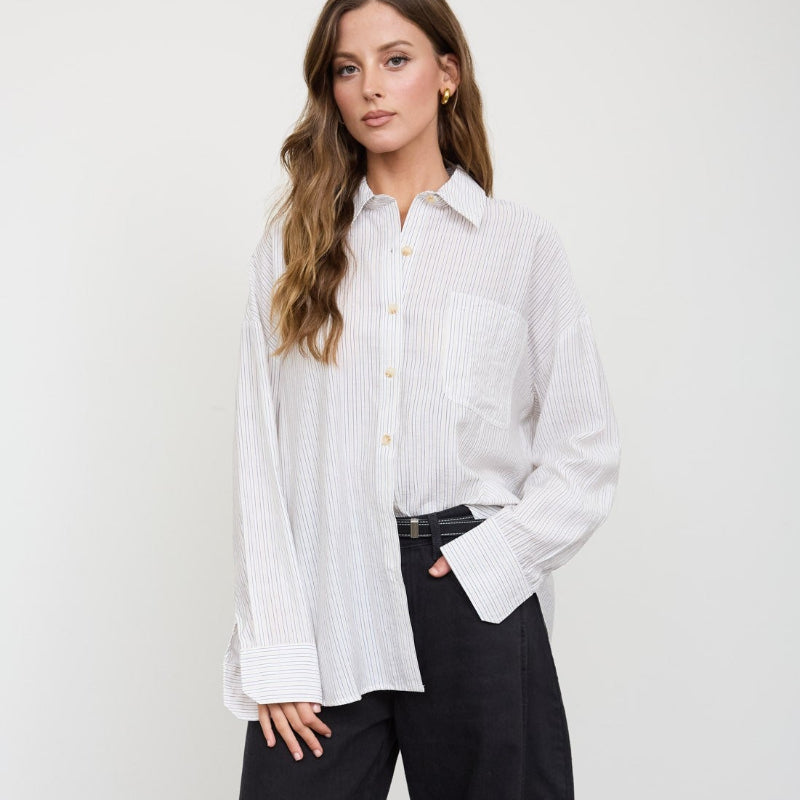 A woman stands against a white wall wearing a white long-sleeve button-up shirt with vertical texture, black wide-leg trousers, and black ankle boots.