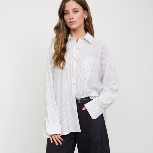 A woman stands against a white wall wearing a white long-sleeve button-up shirt with vertical texture, black wide-leg trousers, and black ankle boots.