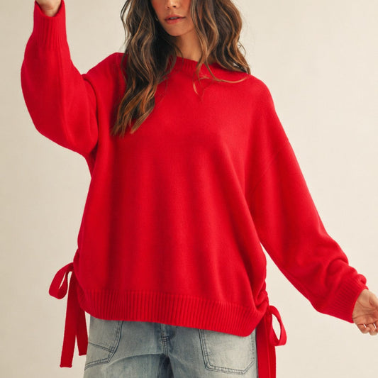 A woman wearing a bright red knit sweater with side ties and light-wash denim shorts, standing against a plain light beige studio background.