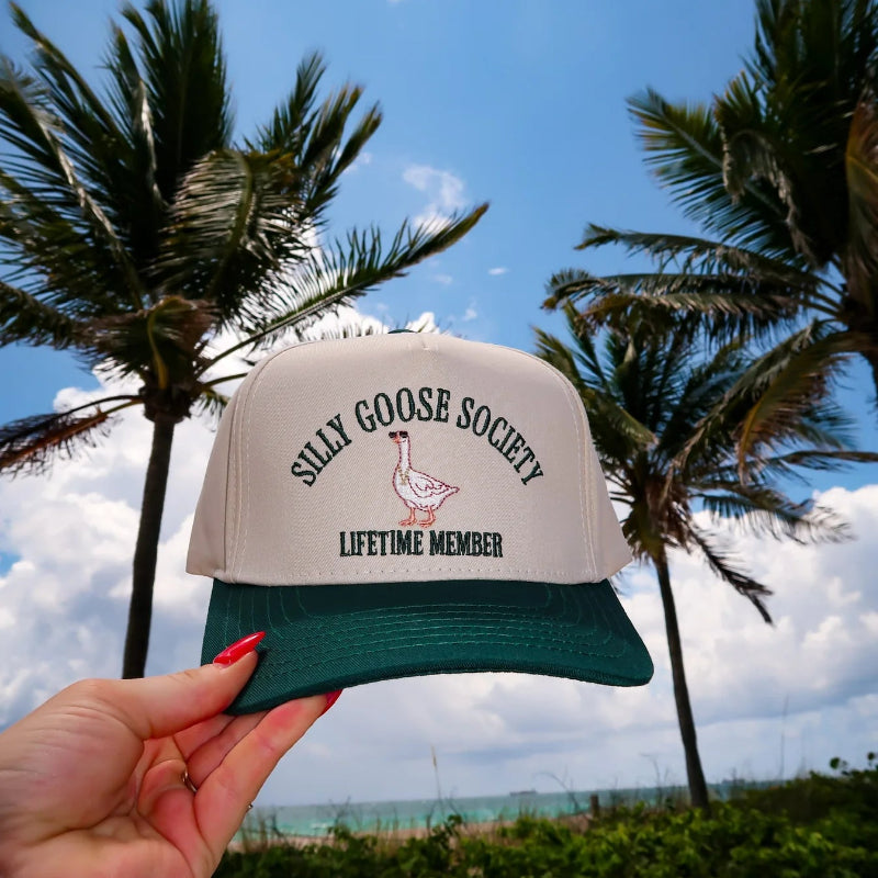 Trucker hat with 'Silly Goose Society Lifetime Member' text held in front of palm trees and blue sky.
