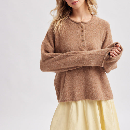 A woman wearing a fuzzy, oversized long-sleeve henley sweater in a warm camel brown, paired with a pleated light yellow skirt, against a plain light gray studio background.