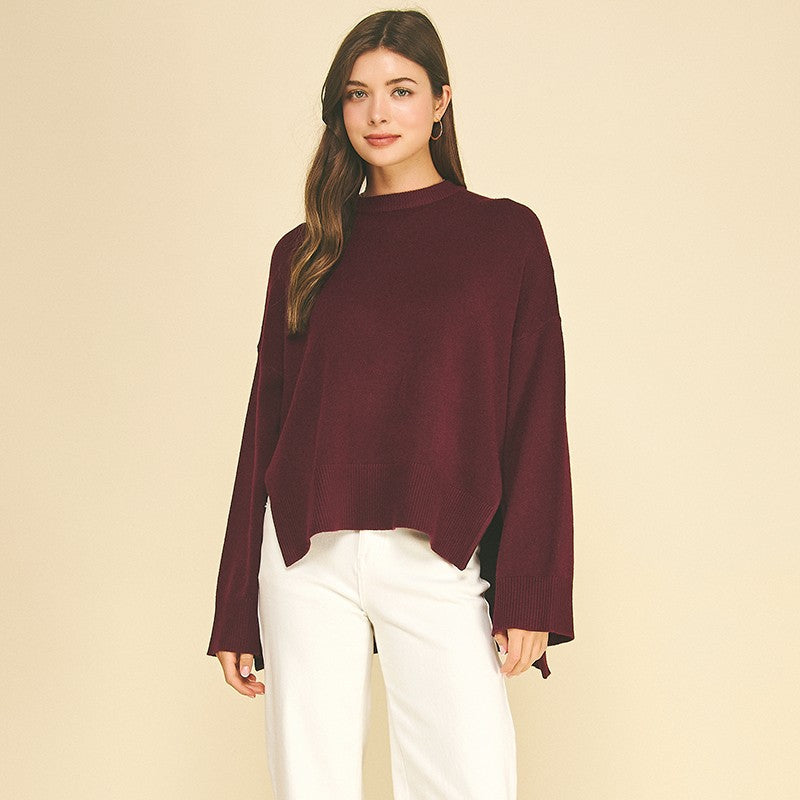 A woman modeling a long-sleeve, cropped mock-neck knit sweater in deep burgundy, paired with white cropped pants and leopard-print flat shoes, against a solid beige studio background.